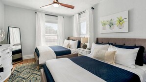 Una habitación de hotel con dos camas, un ventilador de techo, una mesita de noche con lámpara, un aparador y un ventanal grande con cortinas.