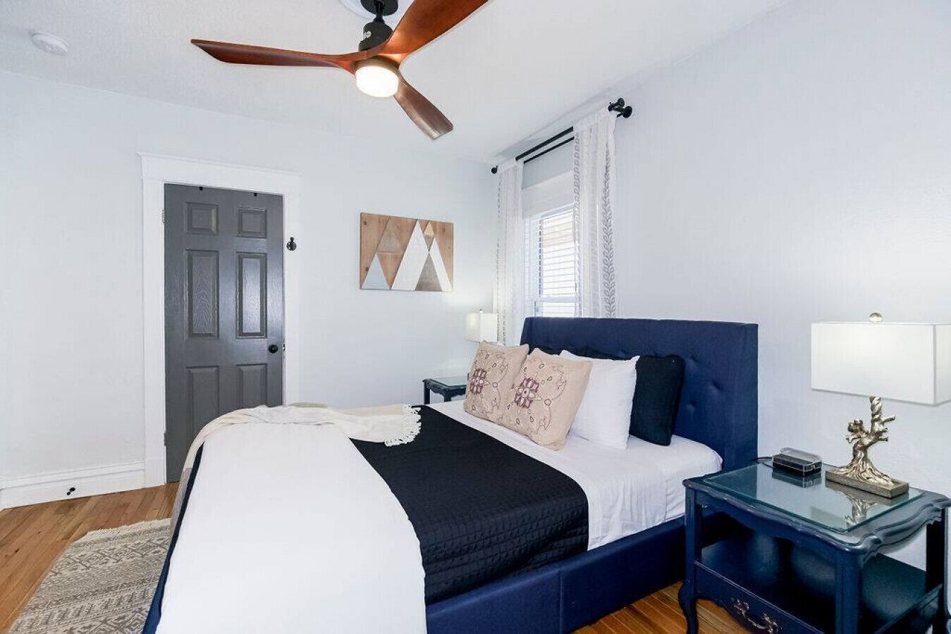 A bedroom with a bed, a ceiling fan, a nightstand, a lamp, a door, and a window with curtains.
