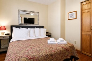 A hotel room with a bed, a bedside table with a lamp, a clock, a mirror, a framed picture on the wall, and a wooden door.