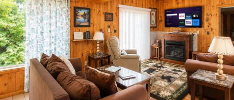 A cozy living room with a sofa, armchair, coffee table, and fireplace. A flat-screen TV is mounted on the wall, and there is a window with curtains.