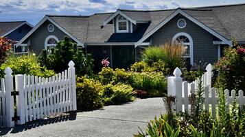 A row of single-story houses with a white picket fence, surrounded by lush greenery and flowering plants.