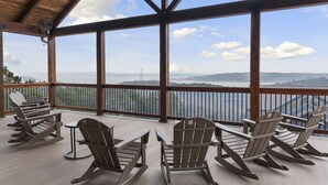 Terraza de madera con sillas Adirondack y una mesa, con vistas a un lago y montañas.