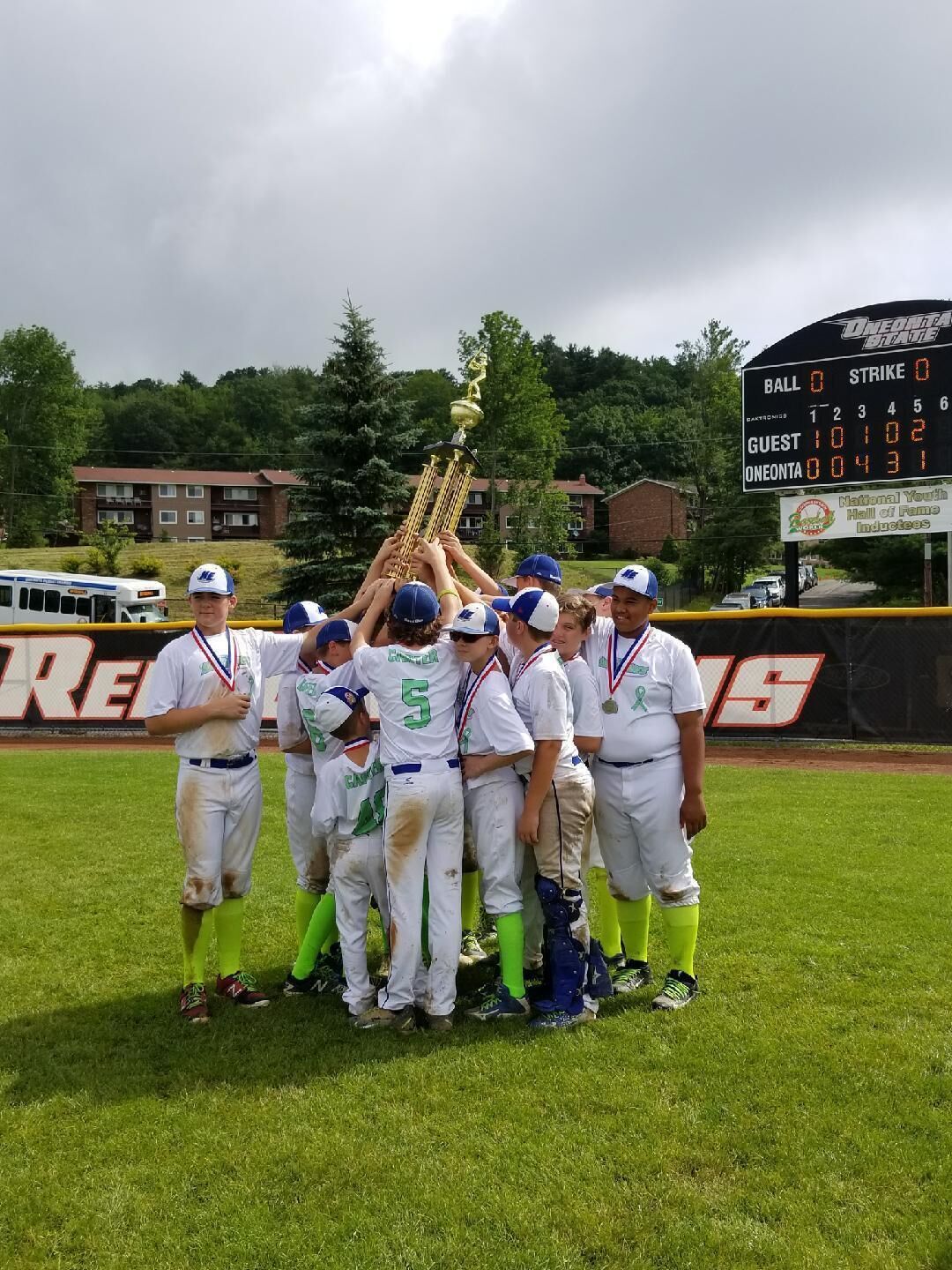 A baseball team huddled around a trophy on a field with a scoreboard in the background.