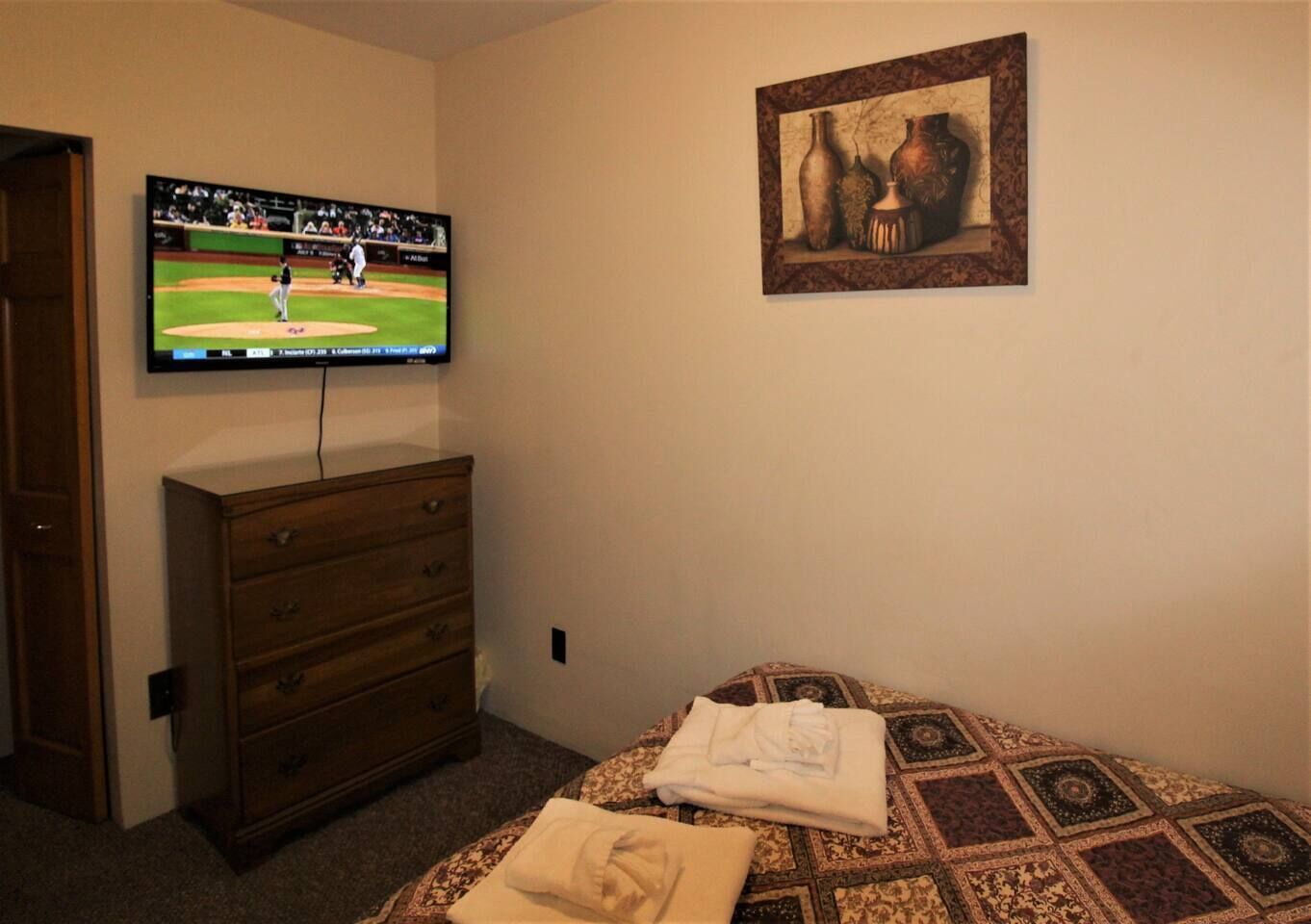 A bedroom with a bed, a dresser, a television displaying a baseball game, and a framed picture on the wall.