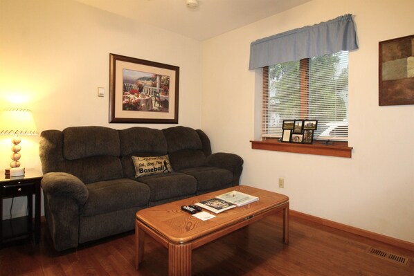 A living room with a sofa, a coffee table, a lamp, a framed picture, and a window with blinds. - Cooperstown Baseball Rentals - Championship #2 (Oneonta)