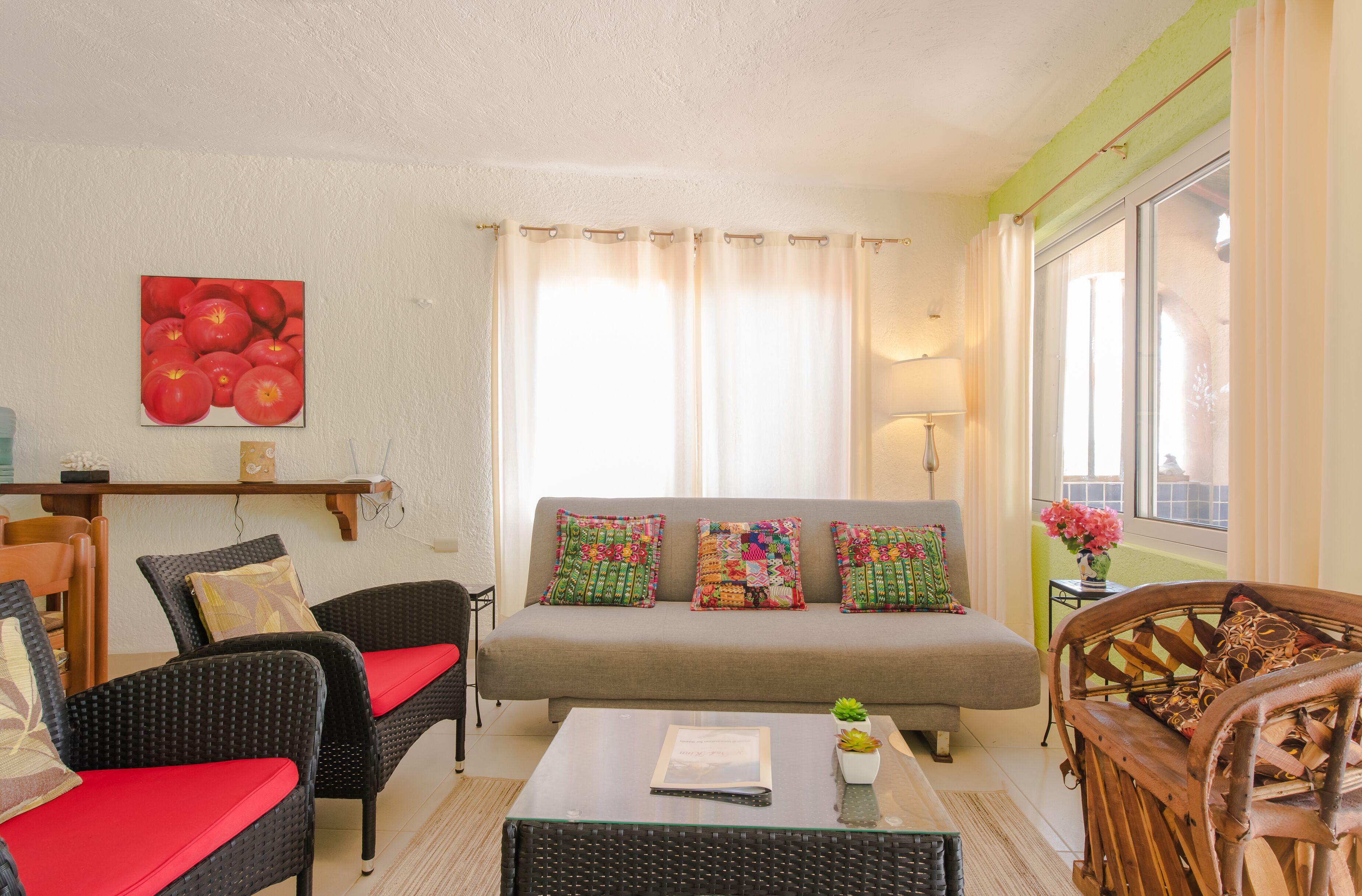 Ein Wohnzimmer mit einer Couch, zwei Korbstühlen mit roten Kissen, einem Glastisch und einem Bild von Tomaten an der Wand.