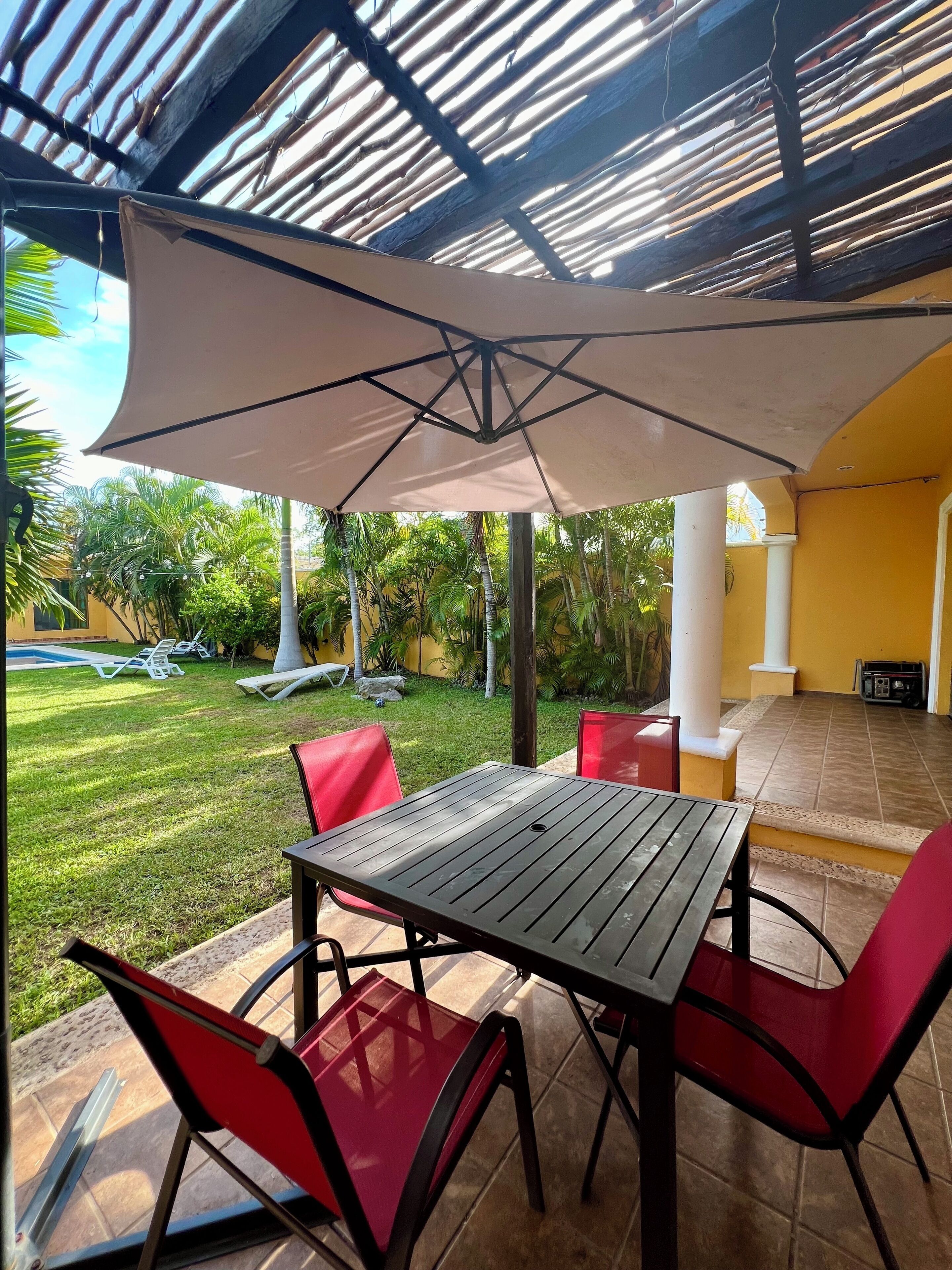 An outdoor patio with a table and chairs, a large umbrella, and a view of a swimming pool and greenery.