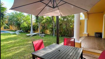 An outdoor patio with a table and chairs, a large umbrella, and a view of a swimming pool and greenery.