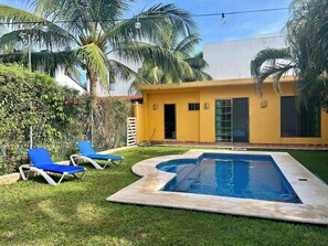 Une propriété résidentielle dotée d’une piscine, de deux fauteuils de jardin bleus et d’une maison jaune.