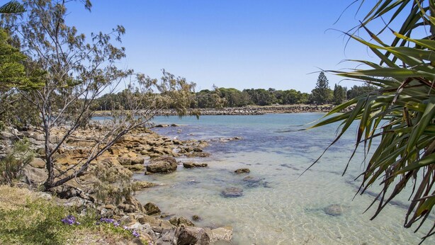 A coastal landscape with clear water, rocky shore, and sparse vegetation.