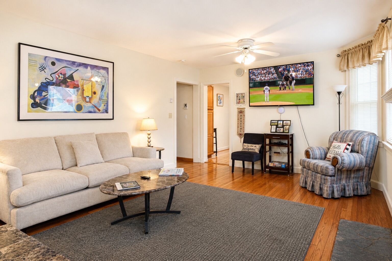 A living room with a sofa, coffee table, armchair, and a television displaying a baseball game.