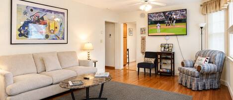 A living room with a sofa, coffee table, armchair, and a television displaying a baseball game.