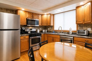 A modern kitchen with stainless steel appliances, wooden cabinets, a granite countertop, and a dining area with a wooden table and chairs. - Cooperstown Baseball Rentals  - Elm-Triple Play #1 (Oneonta)