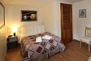 A neatly made bed with a patterned bedspread, a framed picture above it, a wooden door, a chair, and a nightstand with a lamp.