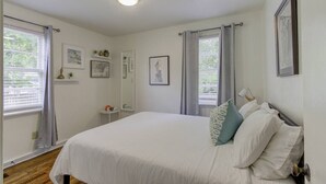 Un dormitorio con una cama grande, dos ventanas con cortinas, una mesita de noche, un espejo y una obra de arte colgada en la pared.