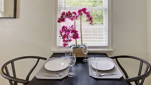 Una mesa de comedor dispuesta con dos platos, cubiertos y copas de vino, con un jarrón de orquídeas rosadas sobre la mesa.