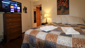 Ein Hotelzimmer mit einem großen Flachbildfernseher, der verschiedene Streaming-Dienste anzeigt, einer Holzkommode, einer gesteppten Bettdecke, einem Blumenbild und einer Nachttischlampe.