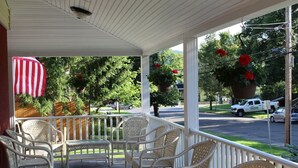 A covered porch with wicker chairs and a table, overlooking a street with parked cars and a truck. - Cooperstown Baseball Rentals - Elm-Triple Play #2R (Oneonta)