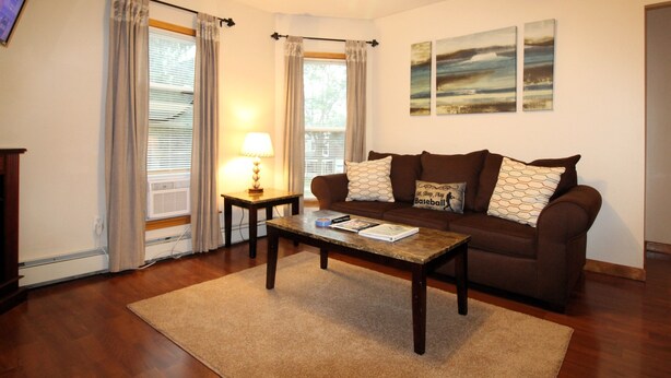 A living room with a brown sofa, a coffee table, a lamp, and a TV. - Cooperstown Baseball Rentals - Elm-Triple Play #2R (Oneonta)
