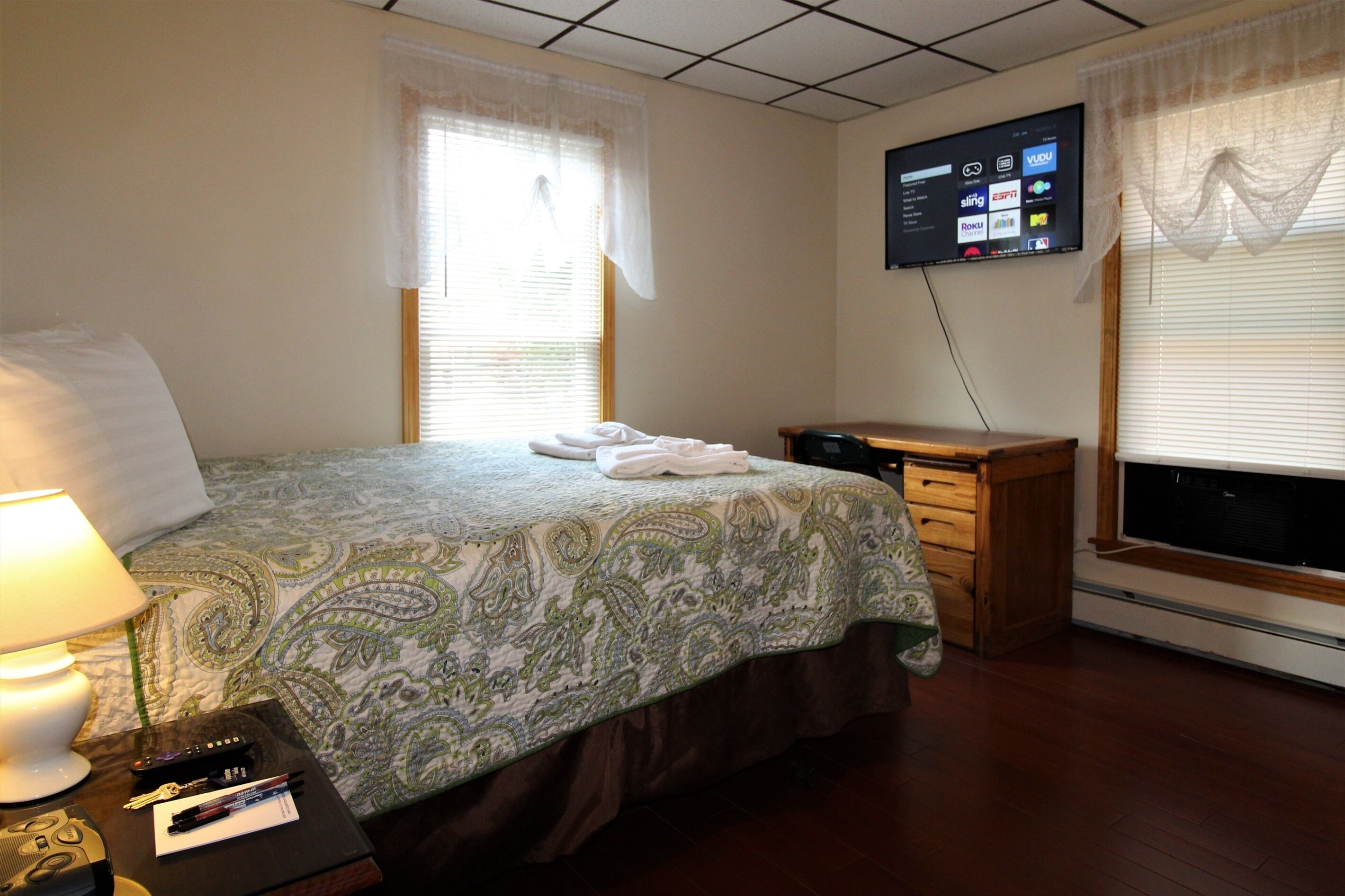 A hotel room with a bed, a desk, a TV mounted on the wall, and a window with blinds.