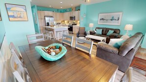 A modern living room with a wooden dining table, a teal accent chair, a brown leather sofa, and a kitchen in the background. - Charming Top-Floor Condo w/ Ocean Views, Pool Access (Tybee Island)