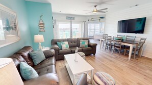 A living room with a brown leather sofa, teal cushions, a white coffee table, and a dining area with wooden furniture. - Charming Top-Floor Condo w/ Ocean Views, Pool Access (Tybee Island)