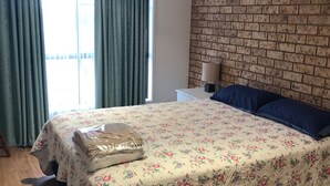 A bedroom with a floral bedspread, a neatly folded blanket, a blue pillow, a brick-patterned wall, and a window with curtains. - 2/36 Sunderland Street, Evans Head (Evans Head)