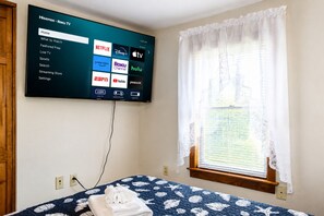 A hotel room with a bed, a TV mounted on the wall displaying a streaming service interface, a window with a lace curtain, and a wooden headboard. - Cooperstown Baseball Rentals - Columbia House (Oneonta)
