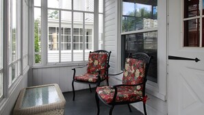 A covered porch with two chairs and a small table, large windows, and a white door.