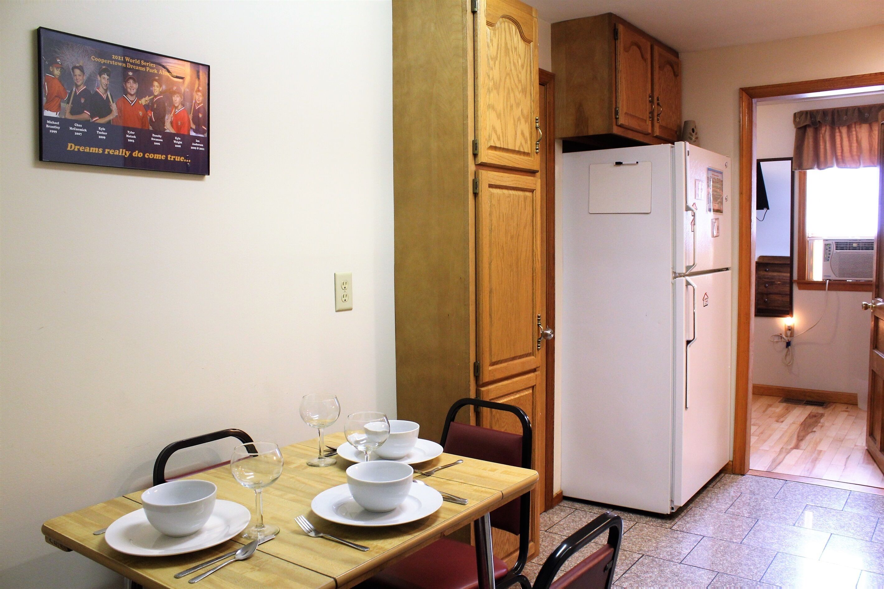 A dining area with a wooden table set for two, a white refrigerator, and a wooden cabinet above the door.
