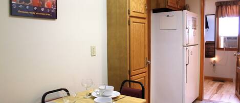 A dining area with a wooden table set for two, a white refrigerator, and a wooden cabinet above the door.