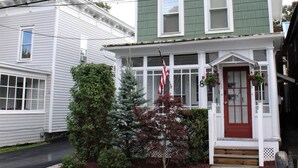 A two-story house with a red front door, white trim, and a green siding. There is a small porch with steps leading to the door, and a Christmas tree with red foliage in front.