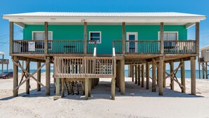 A turquoise two-story beach house with a wooden deck and a balcony, situated on a sandy beach.