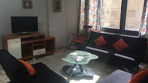 Een woonkamer met een televisie, bank, stoelen, een glazen salontafel en een raam met gordijnen.
