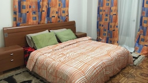 Een slaapkamer met een houten bed, groene en beige kussens, een nachtkastje en behang met een patroon.