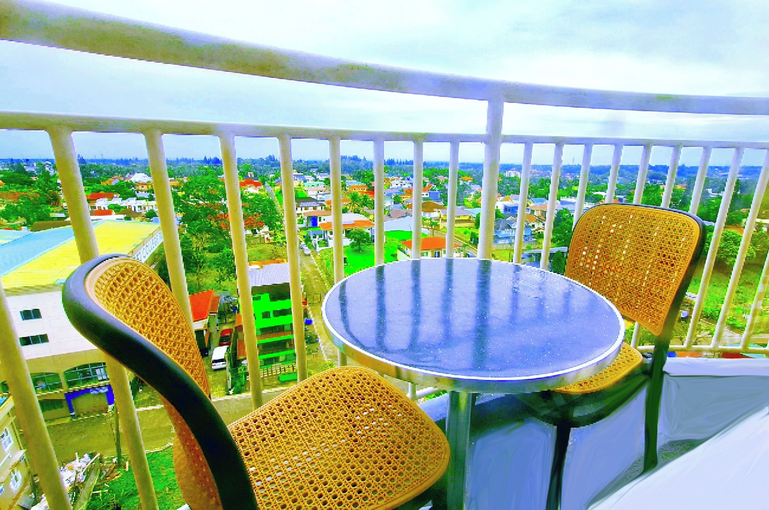 Ein Balkon mit rundem Tisch und zwei Korbstühlen, der auf einen Wohnbereich mit Häusern und Grünflächen blickt.