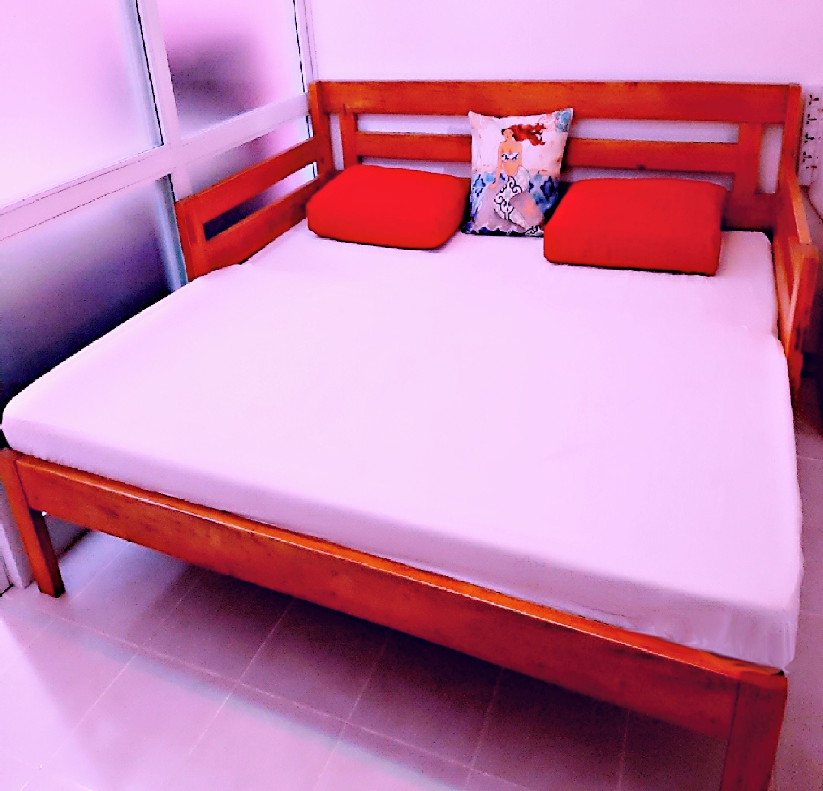 A bed with a wooden frame, red pillows, and a white mattress in a room with white walls and tiled flooring.