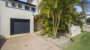 Une maison moderne à deux étages, dotée d’un garage, d’une allée en béton et d’un jardin bien entretenu avec des palmiers.