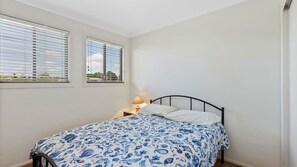 Un dormitorio con cama, dos ventanas con persianas, una mesita de noche con lámpara y vistas a zonas verdes.