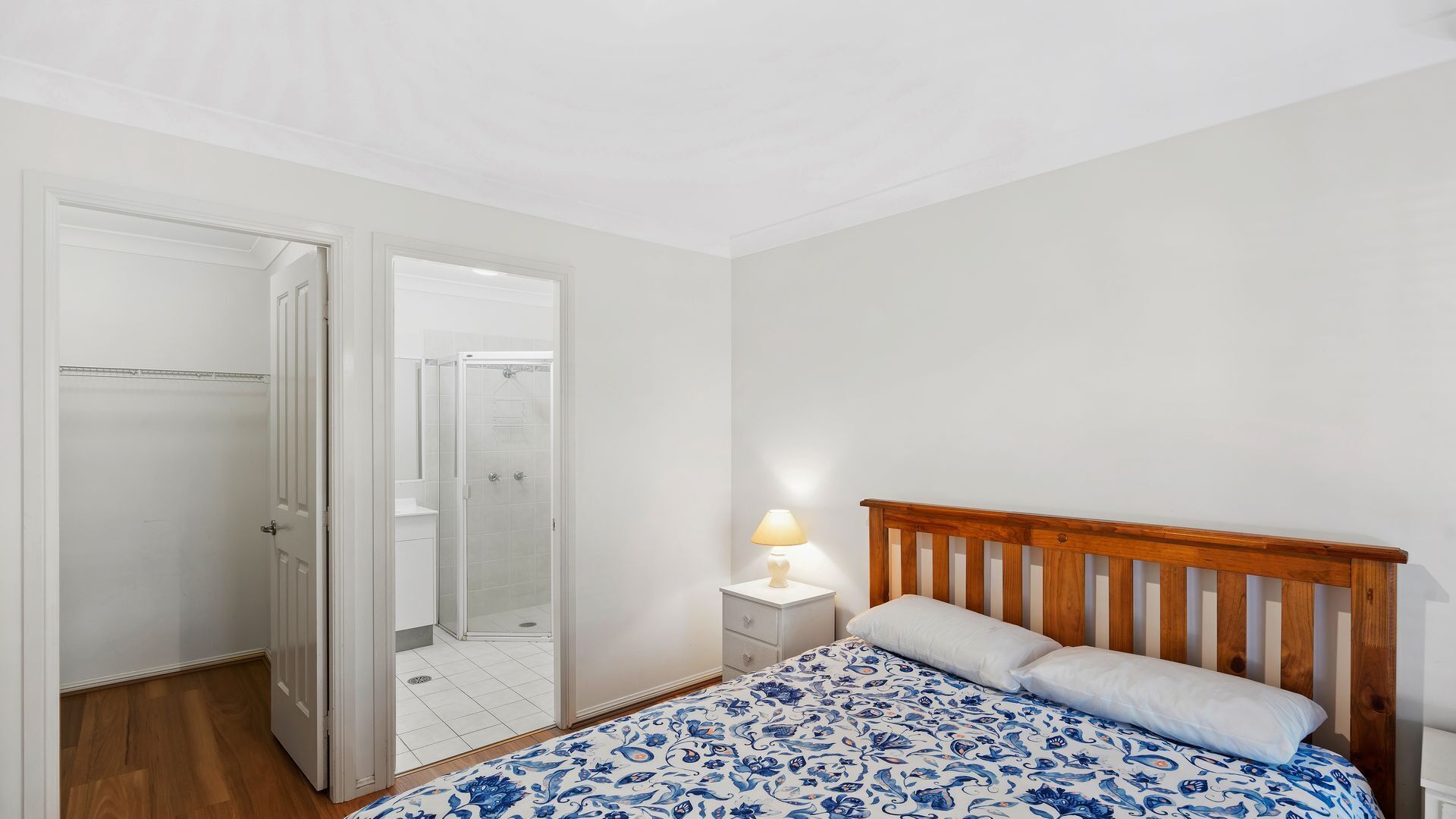 Ein Schlafzimmer mit einem Bett, einem Nachttisch, einer Lampe, einem Holz-Kopfteil und einem Schrank mit Spiegel.