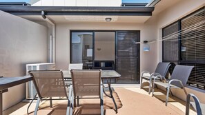 Un patio exterior con una mesa y sillas, una puerta corrediza de vidrio que da a una habitación y una ventana con persianas.