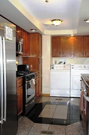 A compact kitchen with wooden cabinets, a stainless steel refrigerator, a microwave, a stove, and a washing machine. - Cooperstown Baseball Rentals - Goose Bay (Portlandville)