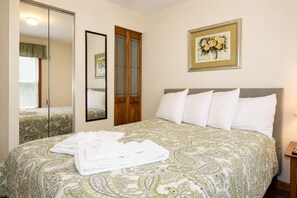 A neatly made bed with a patterned bedspread, a bedside table with a lamp, a mirror, and a framed picture on the wall. - Cooperstown Baseball Rentals - 2nd Baseman (Oneonta)