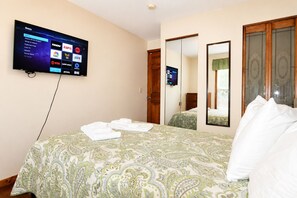 A hotel room with a bed, a TV mounted on the wall displaying channel options, a mirror, and a wardrobe. - Cooperstown Baseball Rentals - 2nd Baseman (Oneonta)