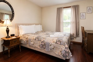 Quarto com cama, mesa de cabeceira com abajur, cômoda e janela com cortinas.