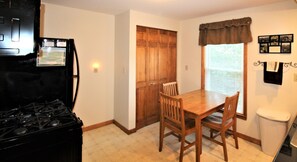 Una cocina con estufa, mesa de madera, sillas, refrigerador y una ventana con persianas.