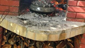 A brick-built outdoor pizza oven with a stone arch, a fire burning inside, and wood logs stored underneath.