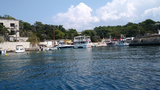 A harbor with boats docked, a stone building, and a backdrop of trees and hills.