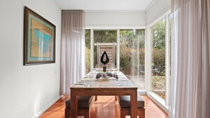 A dining area with a wooden table, chairs, and a view of the outdoors through large windows. - Aireys Oasis (Aireys Inlet)