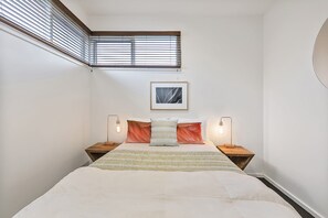 A bedroom with a bed, two bedside tables, a nightstand, a window with blinds, and a framed picture on the wall.